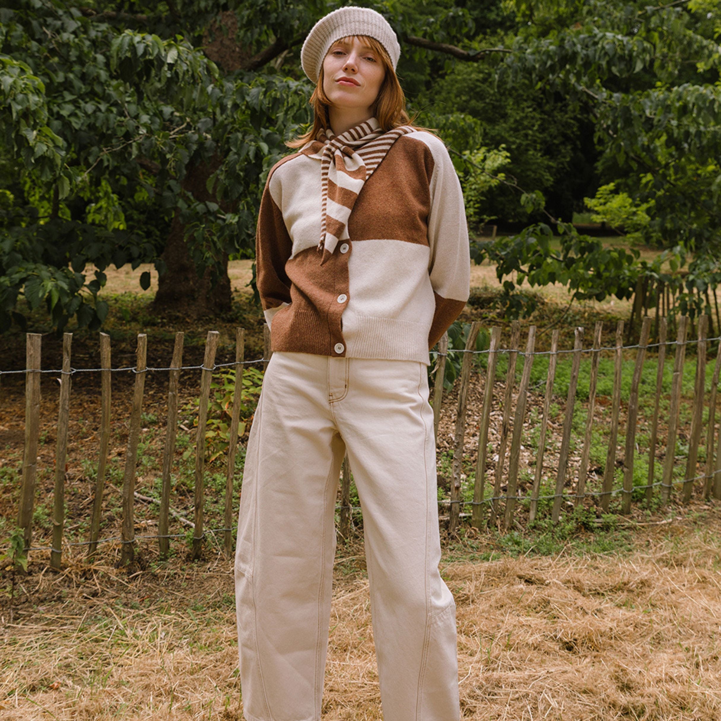 Person wearing a brown and white cardigan with a scarf, standing in a field with trees in the background.