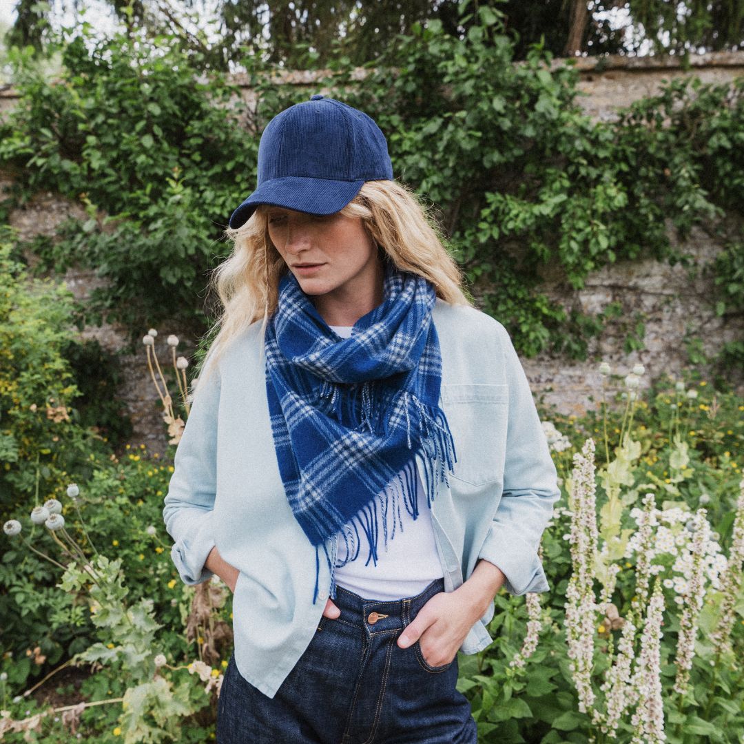 A woman wearing a light blue button-up shirt, dark blue jeans, a navy blue baseball cap, and a blue and white plaid fringed scarf, standing in a garden setting.