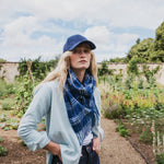 A woman standing in a garden wearing a blue baseball cap, a blue and white plaid scarf, a light blue shirt , and dark jeans.