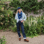 A woman stands in a garden setting wearing a light blue cardigan, a blue patterned scarf, dark blue jeans, a navy blue baseball cap, and brown boots.