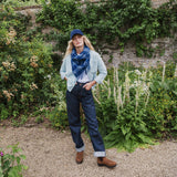 A woman stands in a garden setting wearing a light blue cardigan, a blue patterned scarf, dark blue jeans, a navy blue baseball cap, and brown boots.