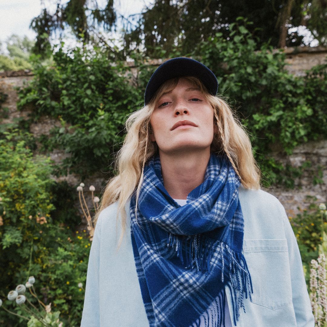 A woman outdoors wearing a blue and white plaid fringed scarf, a light blue cardigan, and a dark navy baseball cap, with green foliage and a stone wall in the background.