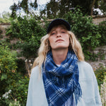 A woman outdoors wearing a blue and white plaid fringed scarf, a light blue cardigan, and a dark navy baseball cap, with green foliage and a stone wall in the background.