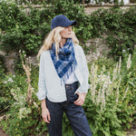 A woman standing in a garden wearing a navy blue plaid fringed scarf, a light gray cardigan, a white t-shirt, dark blue jeans, and a navy baseball cap.