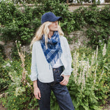 A woman standing in a garden wearing a navy blue plaid fringed scarf, a light gray cardigan, a white t-shirt, dark blue jeans, and a navy baseball cap.
