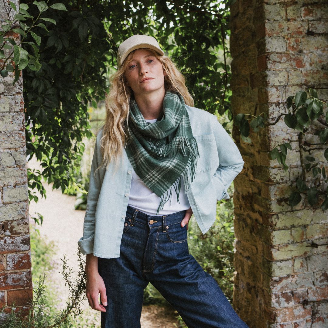 A woman wearing a green and gray plaid fringed scarf, a light blue denim shirt, a white t-shirt, and dark blue jeans, standing outdoors between brick pillars and green foliage.