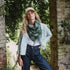 A woman wearing a green and gray plaid fringed scarf, a light blue denim shirt, a white t-shirt, and dark blue jeans, standing outdoors between brick pillars and green foliage.