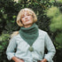 a woman standing beside a bush wearing a green cable knit scarf.