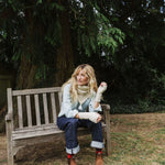 A blonde lady sat on a bench wearing a cream cable knit scarf.