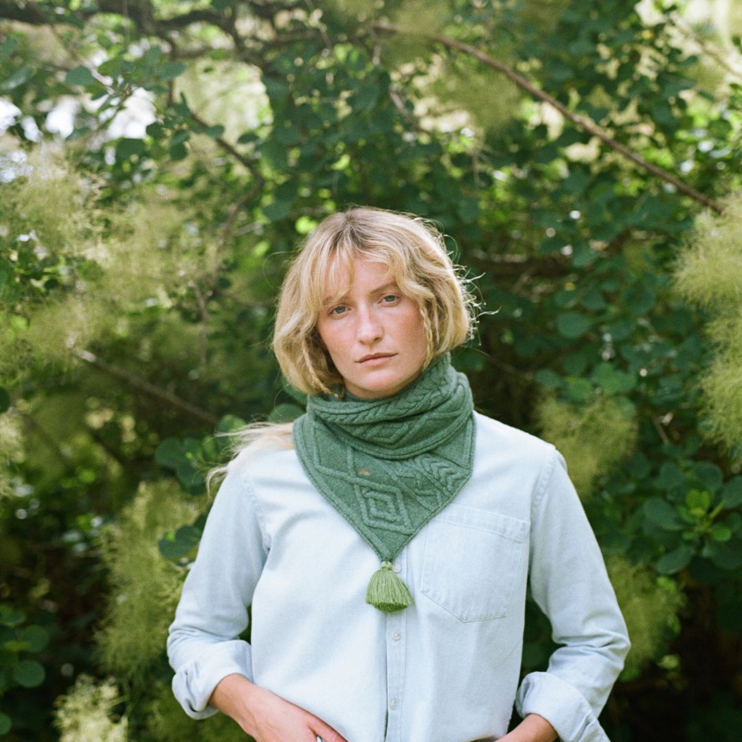 A lady with blonde hair standing in front of a tree wearing a green scrarf.