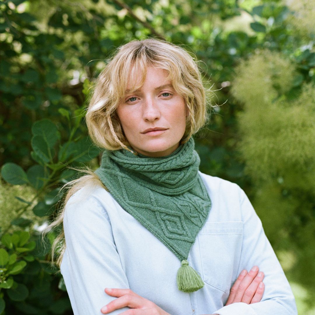 A smiling lady standing with rms folded wearing a green cable knit scarf.