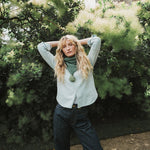 A lady wearing a green cable knit scarf with her hands above her head standing infront of a tree.