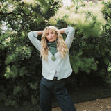 A lady wearing a green cable knit scarf with her hands above her head standing infront of a tree.