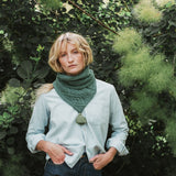 woman standing by a whispy bush wearing a soft green cable knit scarf and blue jeans.