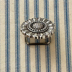 An ornate silver-toned metal scarf slider with a floral sunburst design and a rectangular pin base, resting on a blue and cream striped fabric background.