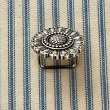 An ornate silver-toned metal scarf slider with a floral sunburst design and a rectangular pin base, resting on a blue and cream striped fabric background.