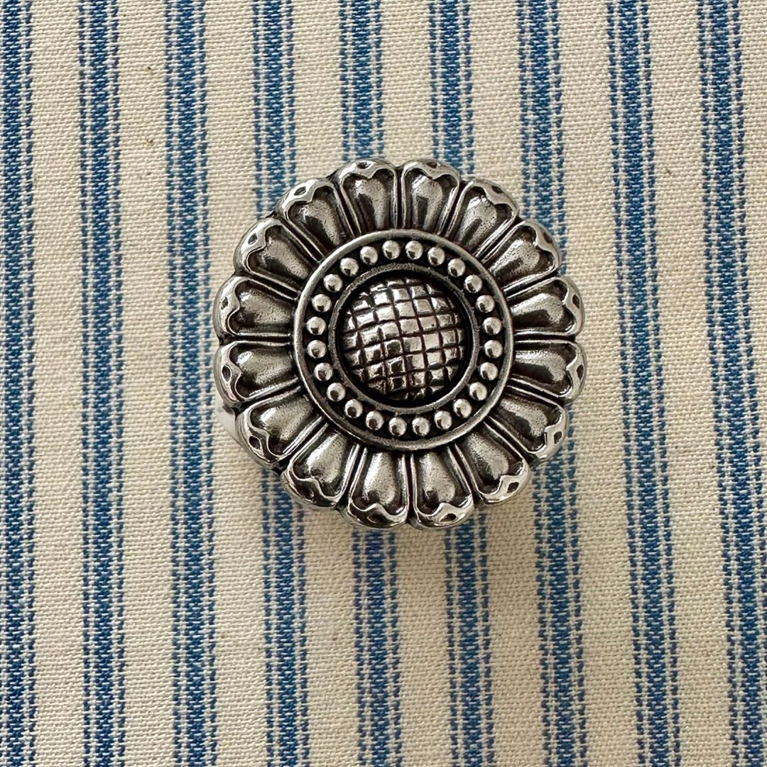 An ornate round silver metal scarf slider with a floral petal design and textured center, placed on a blue and cream striped fabric background.
