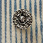 An ornate round silver metal scarf slider with a floral petal design and textured center, placed on a blue and cream striped fabric background.