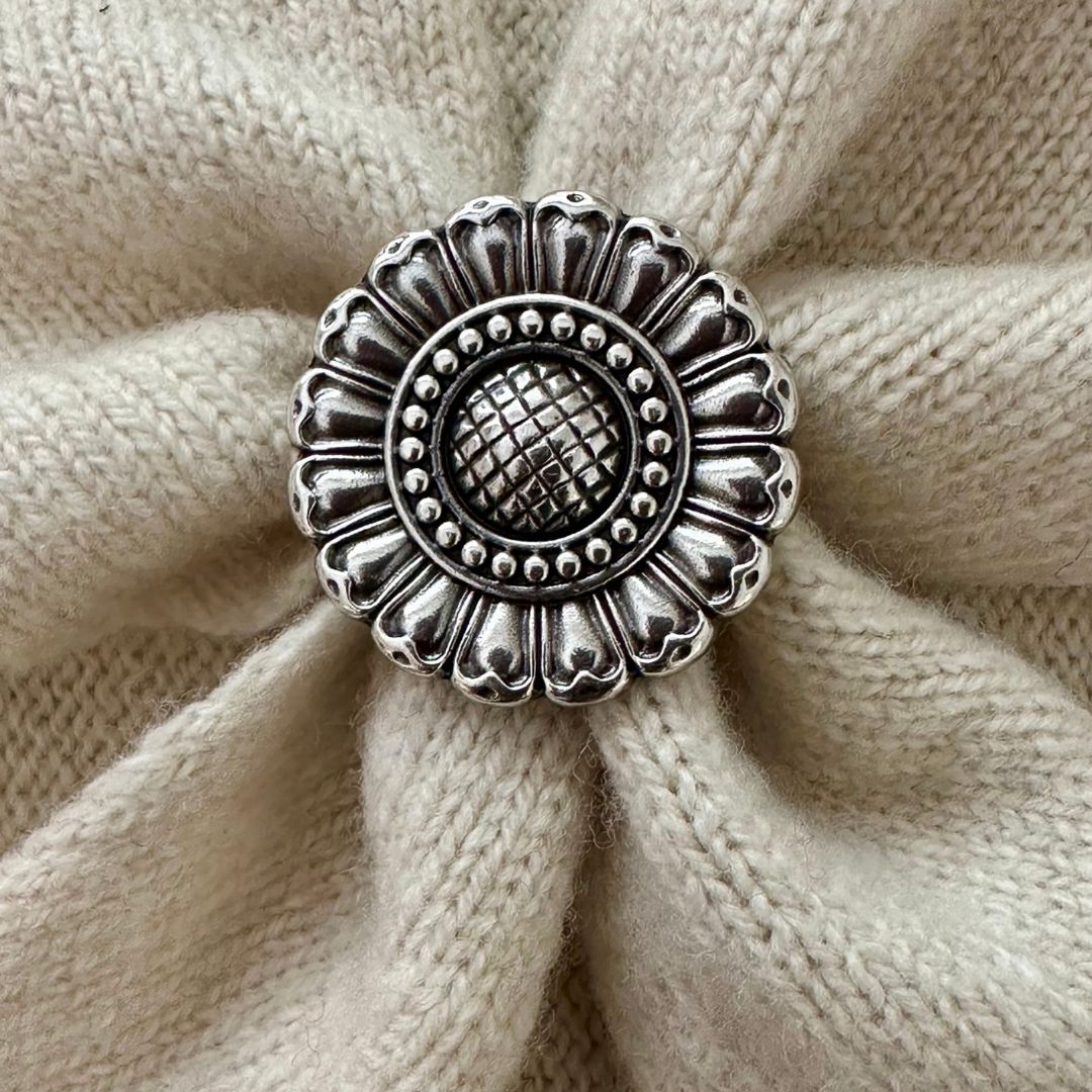 A round silver-toned metal scarf slider with an ornate sunflower-like pattern, placed on a textured beige knit fabric background.