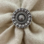 A round silver-toned metal scarf slider with an ornate sunflower-like pattern, placed on a textured beige knit fabric background.