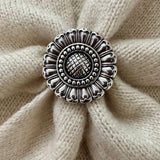 A round silver-toned metal scarf slider with an ornate sunflower-like pattern, placed on a textured beige knit fabric background.