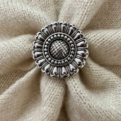 A round silver-toned metal scarf slider with an ornate sunflower-like pattern, placed on a textured beige knit fabric background.