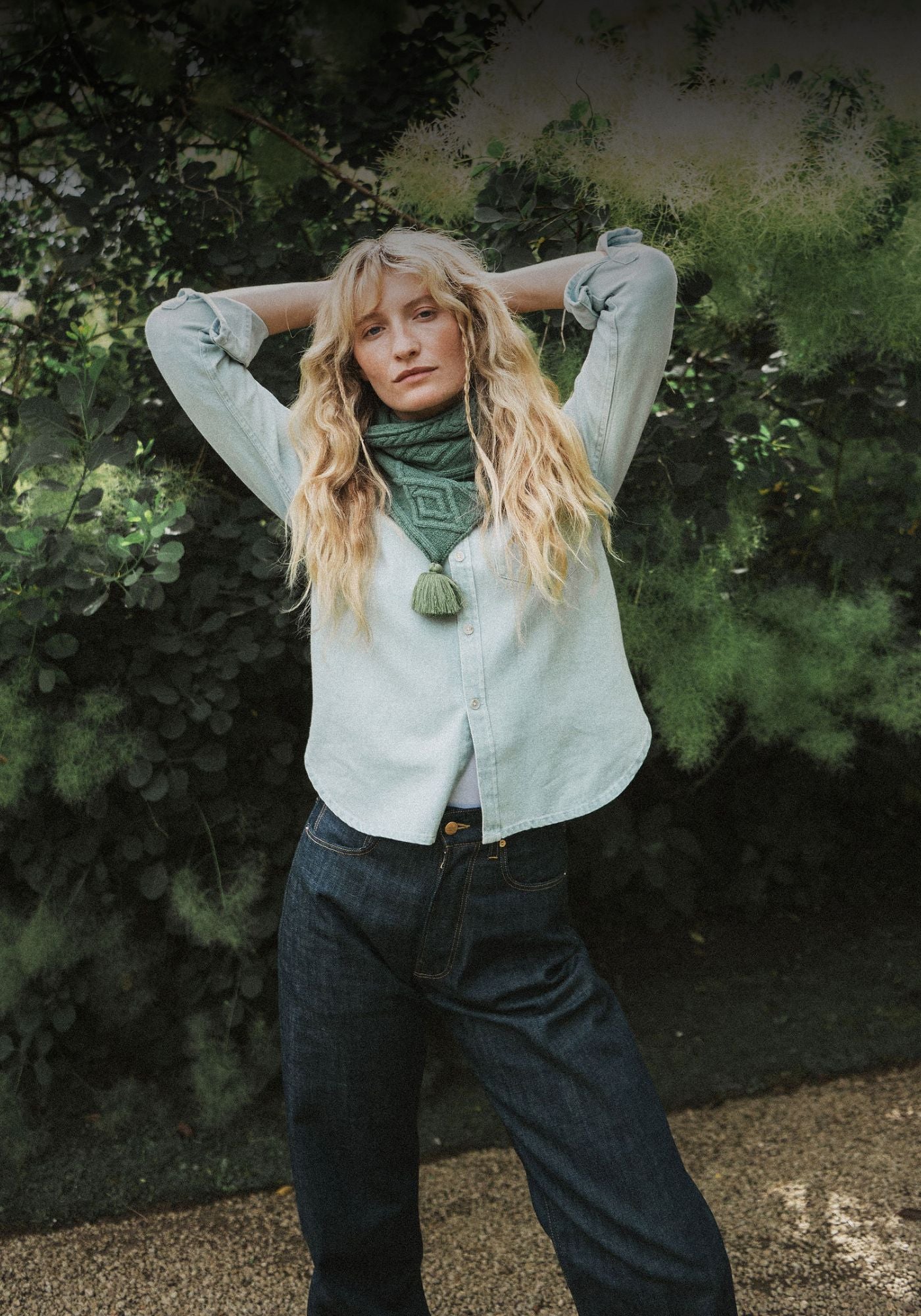 A woman with long blonde hair poses outdoors against a backdrop of green foliage, wearing a light sage green button-up shirt, a dark green patterned knitted swisted snood, and dark blue jeans.