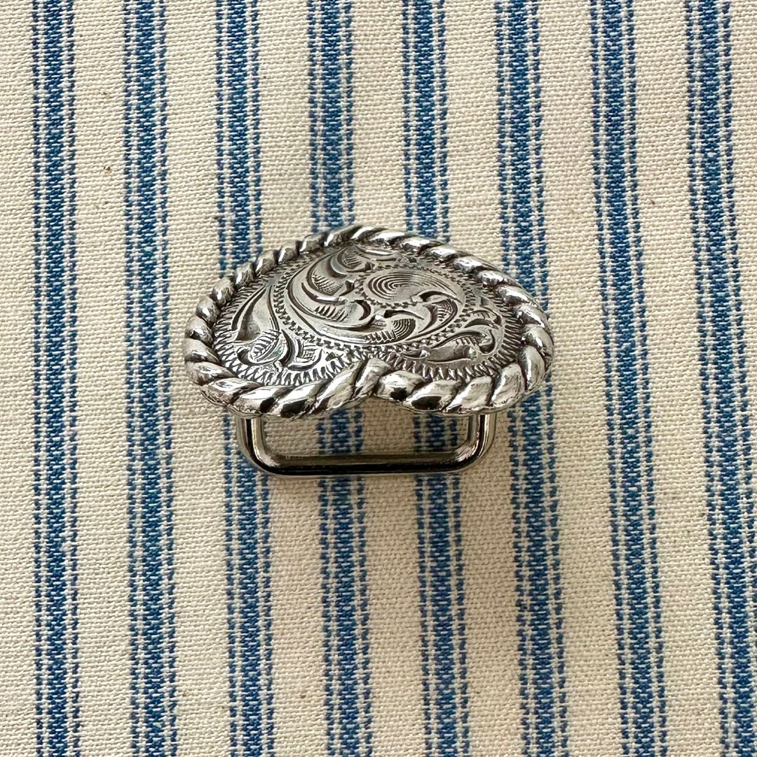 A silver-toned scarf slider with an engraved floral pattern and twisted rope border, placed on a blue and cream striped fabric background.