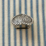 A silver-toned scarf slider with an engraved floral pattern and twisted rope border, placed on a blue and cream striped fabric background.