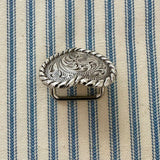 A silver-toned scarf slider with an engraved floral pattern and twisted rope border, placed on a blue and cream striped fabric background.