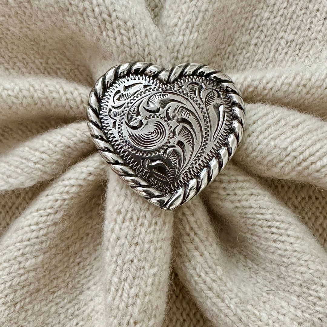 A heart-shaped silver-toned metal brooch with intricate scrollwork and a twisted border, displayed on a textured cream knit fabric.