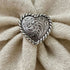 A heart-shaped silver-toned metal brooch with intricate scrollwork and a twisted border, displayed on a textured cream knit fabric.