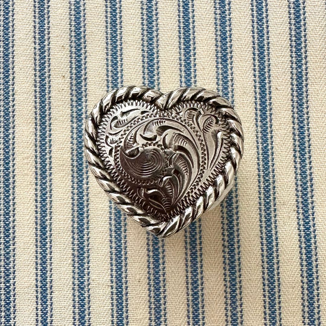 A heart-shaped silver-toned metal scarf slider with intricate floral engravings and a twisted rope border, displayed on a blue and cream striped fabric background.