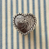 A heart-shaped silver-toned metal scarf slider with intricate floral engravings and a twisted rope border, displayed on a blue and cream striped fabric background.