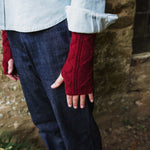 A person wearing dark blue jeans and a light-colored shirt, showing off a pair of deep red cable-knit fingerless gloves.