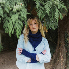 A woman standing outdoors under pine tree branches, wearing a light blue button-up shirt, a dark navy knit scarf, and matching navy knit wrist warmers.