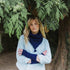 A woman standing outdoors under pine tree branches, wearing a light blue button-up shirt, a dark navy knit scarf, and matching navy knit wrist warmers.