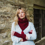 A woman with blonde hair stands against a rustic stone wall, wearing a light blue shirt, a deep red cable-knit scarf, and matching red fingerless knit gloves.
