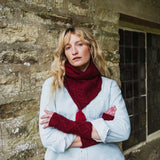 A woman with blonde hair stands against a rustic stone wall, wearing a light blue shirt, a deep red cable-knit scarf, and matching red fingerless knit gloves.