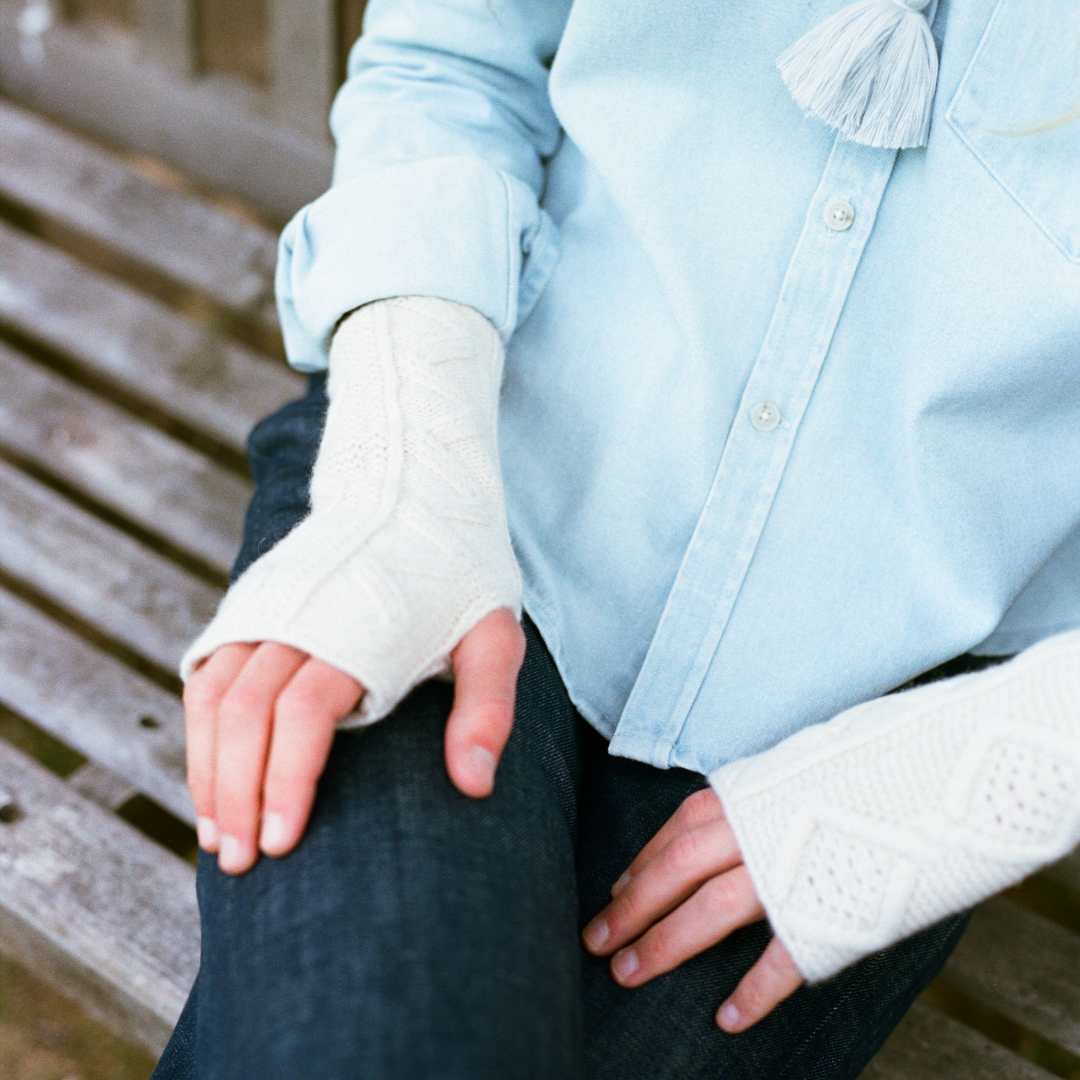 A close-up of a person sitting on a wooden bench, wearing a light blue button-up shirt, dark jeans, and cream-colored fingerless knit  wrist warmers.with cable-knit detailing.