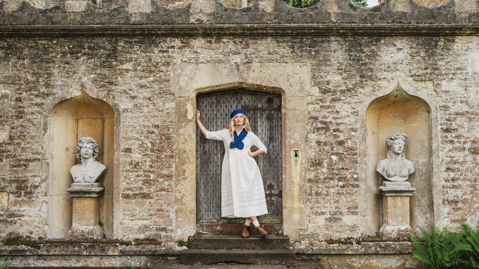 lady standing in a doorway of a cotswold stone building wearing a white dress, blue beret and a blue scarf.