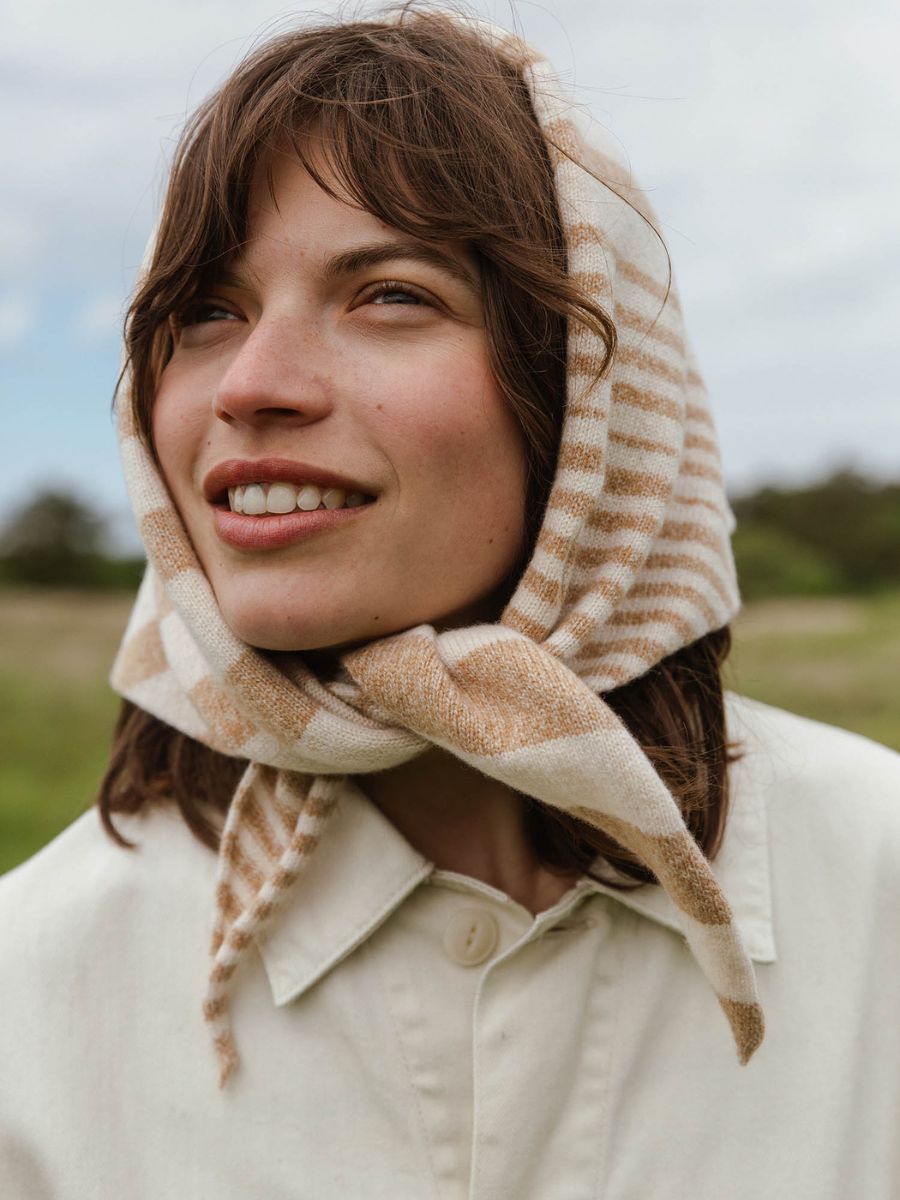 A woman outdoors wearing a cream and tan striped knited scarf on her head, tied under her chin, paired with a light cream button-up shirt.