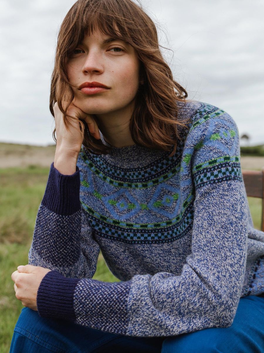 A woman wearing a blue and navy Fair Isle knit sweater with green accents, sitting outdoors in a grassy field under an overcast sky.