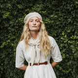 A woman standing outdoors, wearing a cream knit beret, a white dress, and a cream neck scarf. The background features a lush green ivy-covered wall with dappled sunlight.