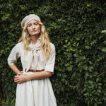 A woman standing outdoors, wearing a cream knit beret, a white dress, and a cream neck scarf. The background features a lush green ivy-covered wall with dappled sunlight.
