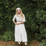 A woman stands outdoors in front of a dense green hedge, wearing a long white linen dress with a tie-neck, a beige beret, a brown belt, white socks, and brown shoes.