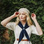 A woman wearing a cream-colored knit beret, a cream dress, and a navy blue neck scarf with a silver button, posing in front of a green ivy-covered wall.