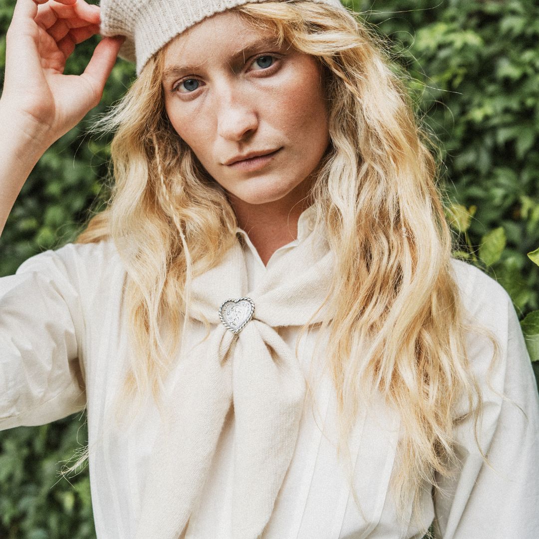 A woman standing outdoors, wearing a cream knit beret, a white dress, and a cream neck scarf. The background features a lush green ivy-covered wall with dappled sunlight.