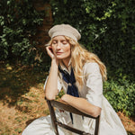 A woman sitting outdoors on a wooden chair, wearing a cream knit beret, a white dress, and a navy blue neck scarf. The background features a lush green ivy-covered wall with dappled sunlight.