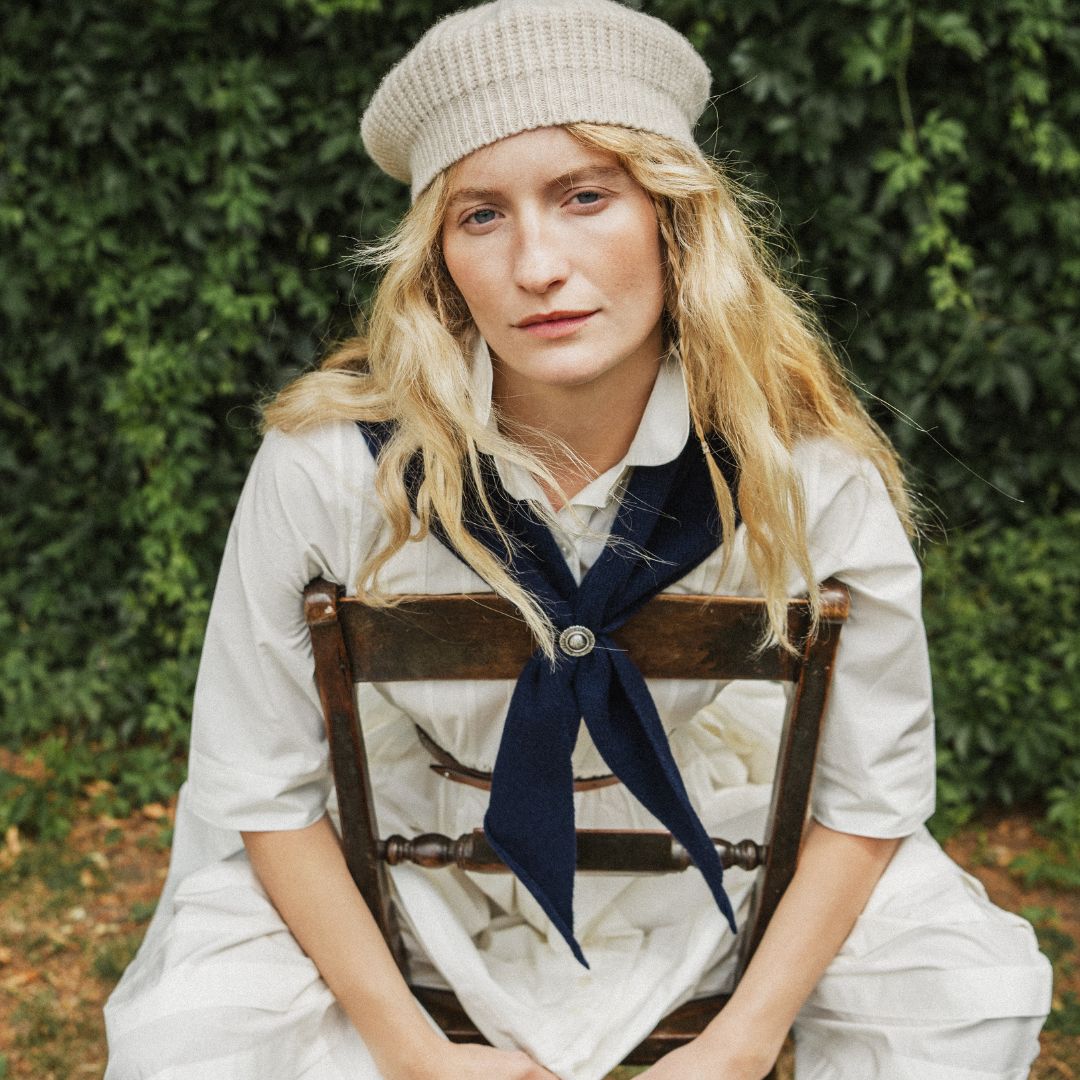 A woman with blonde hair sits on a wooden chair outdoors, wearing a cream knit beret, a white short-sleeve collared shirt with a navy necktie, and white trousers, with a dense green hedge behind her.